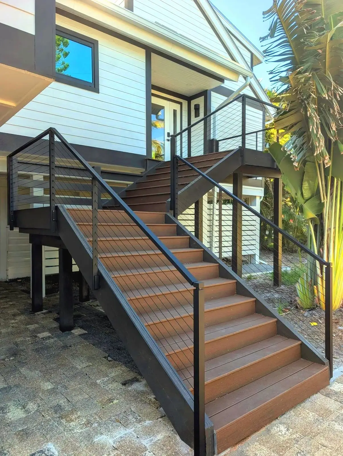 Black cable railing on a wood exterior staircase up to a white stilt house, paver landing and palms