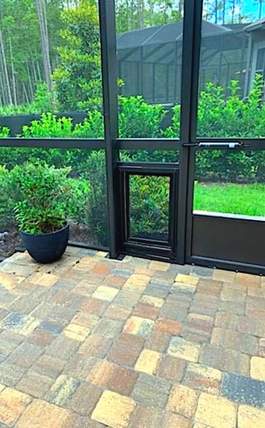 Screened lanai with black aluminum framing, pet door panel and paver floor, screened pool enclosure and woods beyond