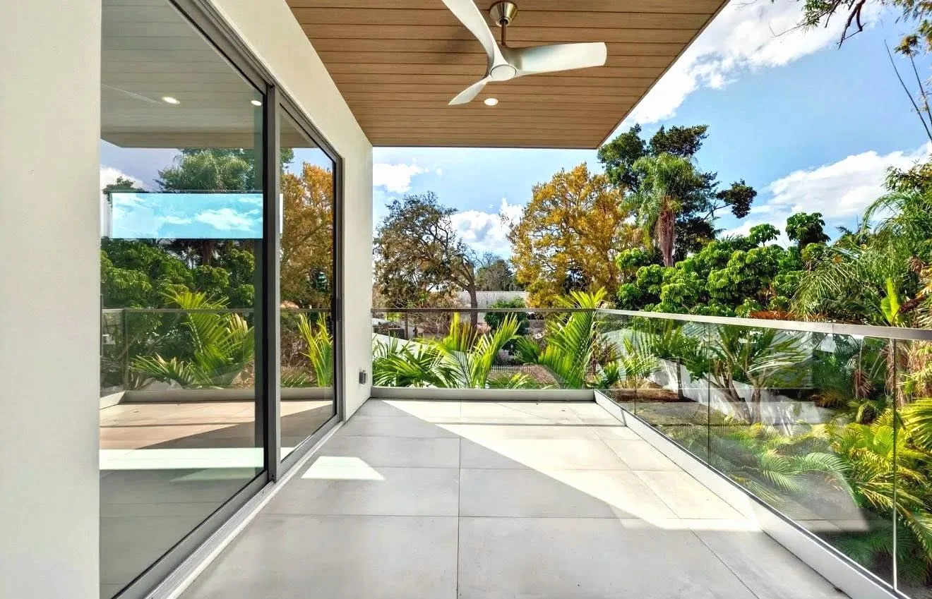 Frameless glass railing on a covered second-floor balcony with a wood ceiling and ceiling fan, trees beyond