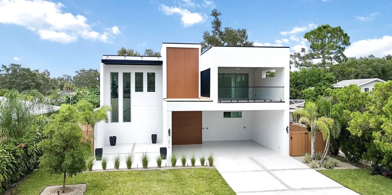 White two-story modern home with a black overhang above tall windows, wood-look cladding accents, glass balcony railing and palms
