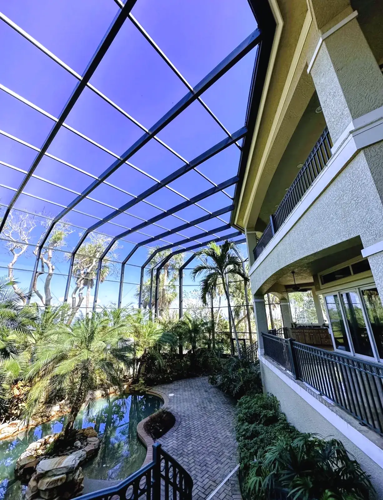 Two story stucco home with black balcony railings inside a tall dark framed screen enclosure over a lagoon pool