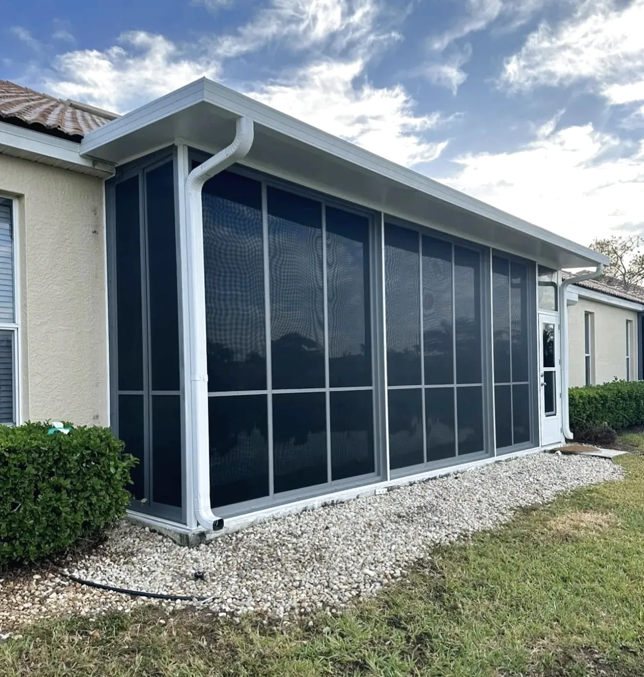 Screened lanai with a white aluminum roof, gutter and downspout on a beige stucco home, gravel border below