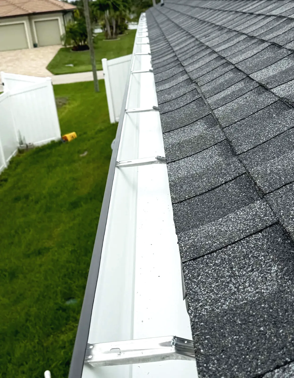Close view along a white seamless gutter run fitted to the edge of a grey shingle roof above a lawn.