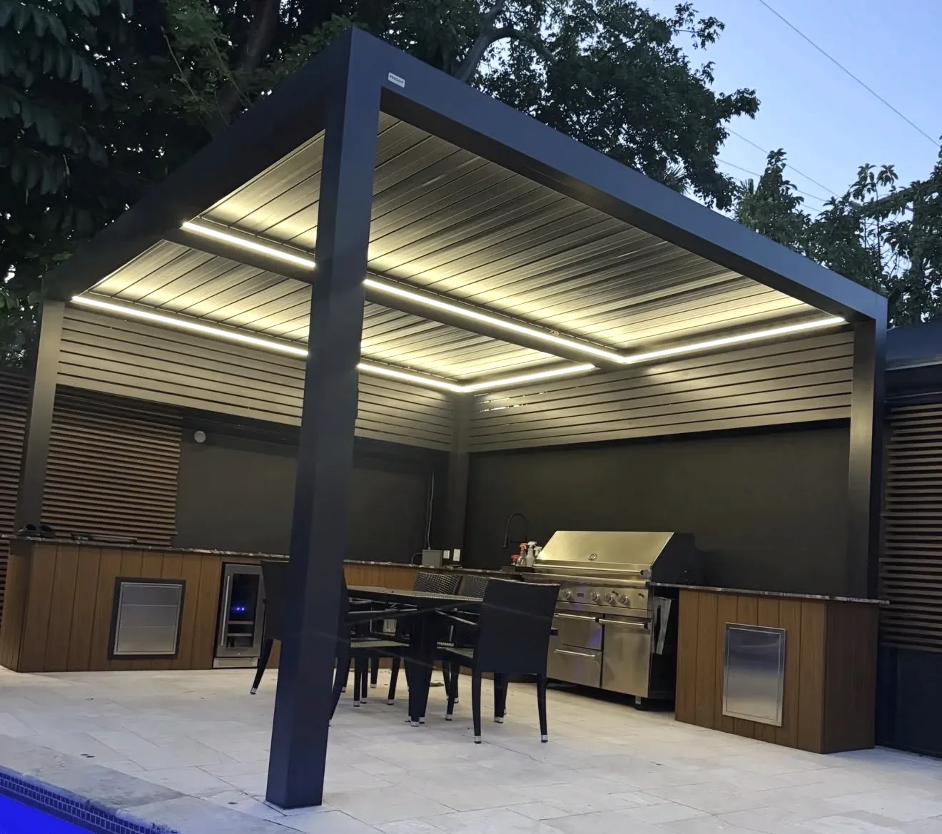 Grey aluminum louvered pergola with strip lighting over an outdoor kitchen, grill and dining set at dusk