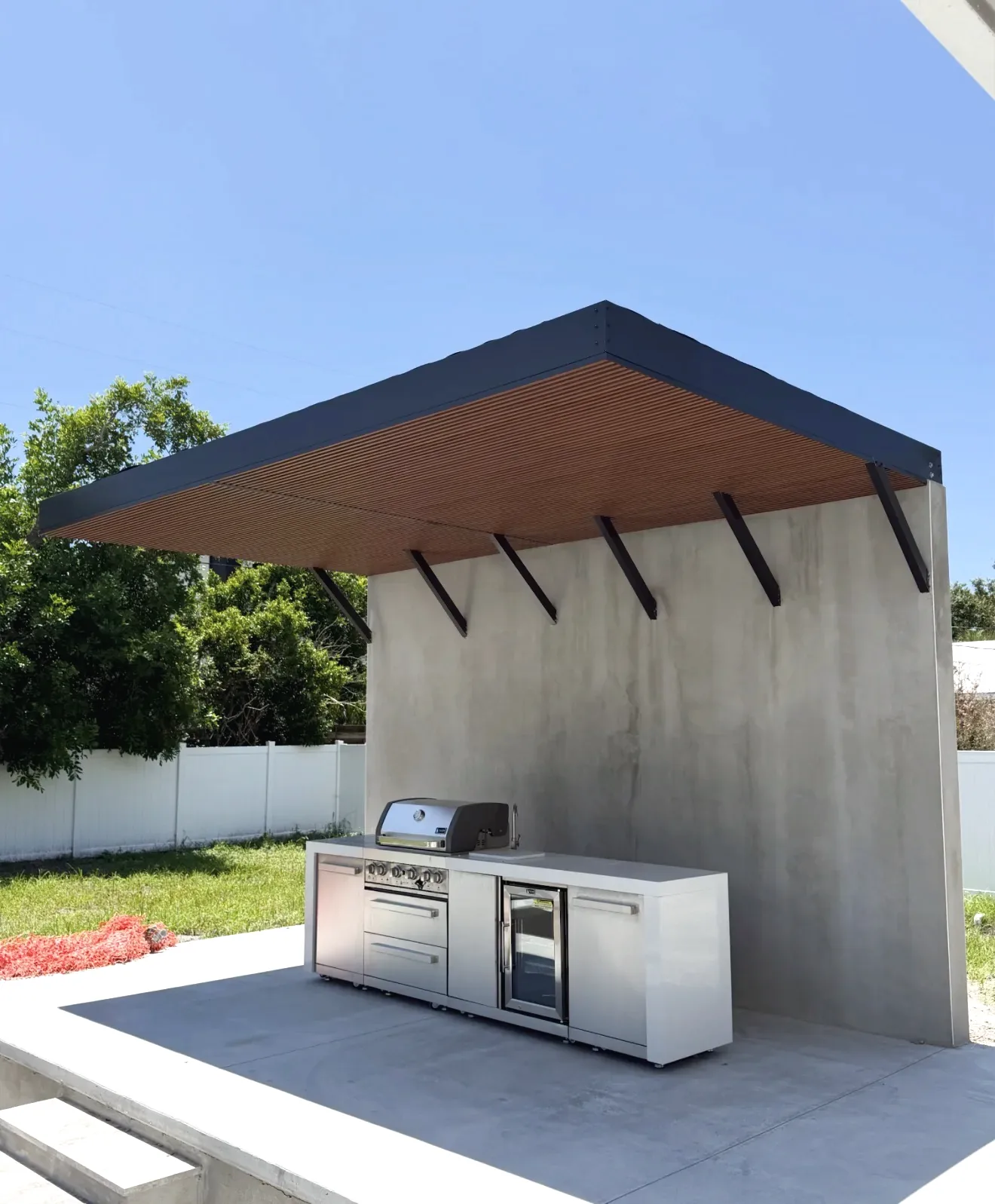 Dark aluminum flat roof canopy with wood grain slat underside, cantilevered over a stainless outdoor kitchen on a concrete patio.