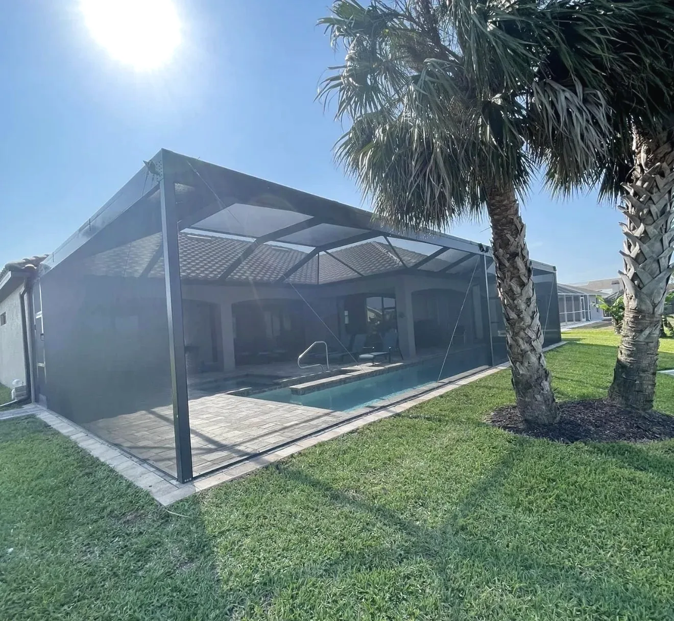 Bronze framed pool cage with dark screening enclosing a pool and paver deck beside a tile roof home and palms.