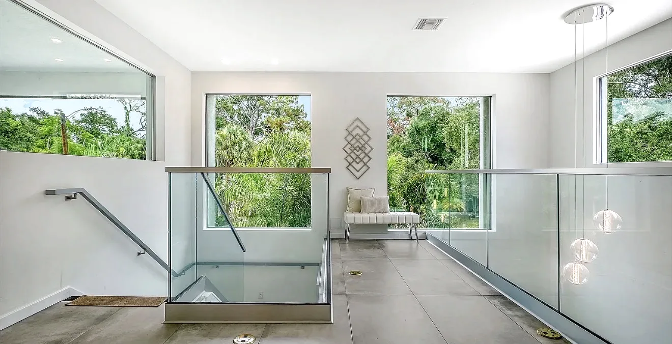 Frameless glass railings with brushed metal caps around a stairwell opening, with a wall-mounted handrail and large windows