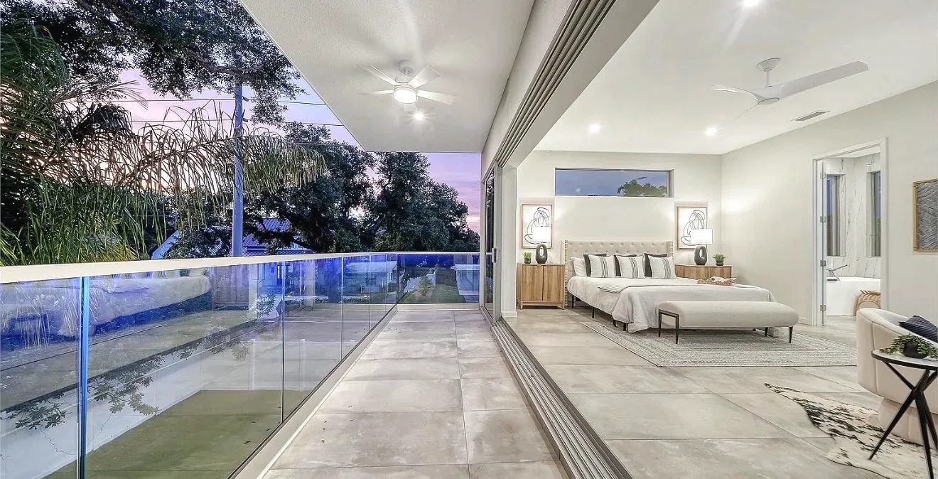 Frameless glass railing along a covered balcony at dusk, sliding glass wall open to a bedroom, pool visible below