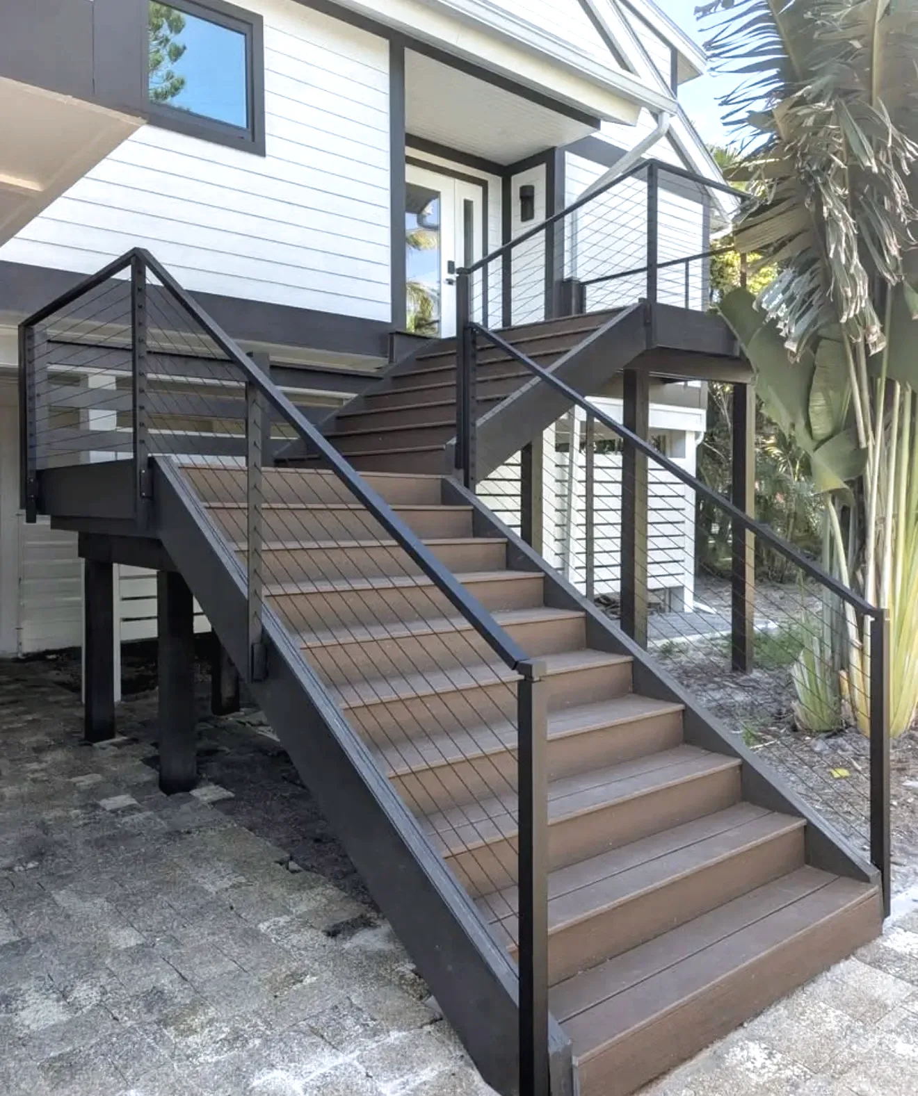 Black cable railing along composite deck stairs rising to an elevated white lap siding home with palms behind