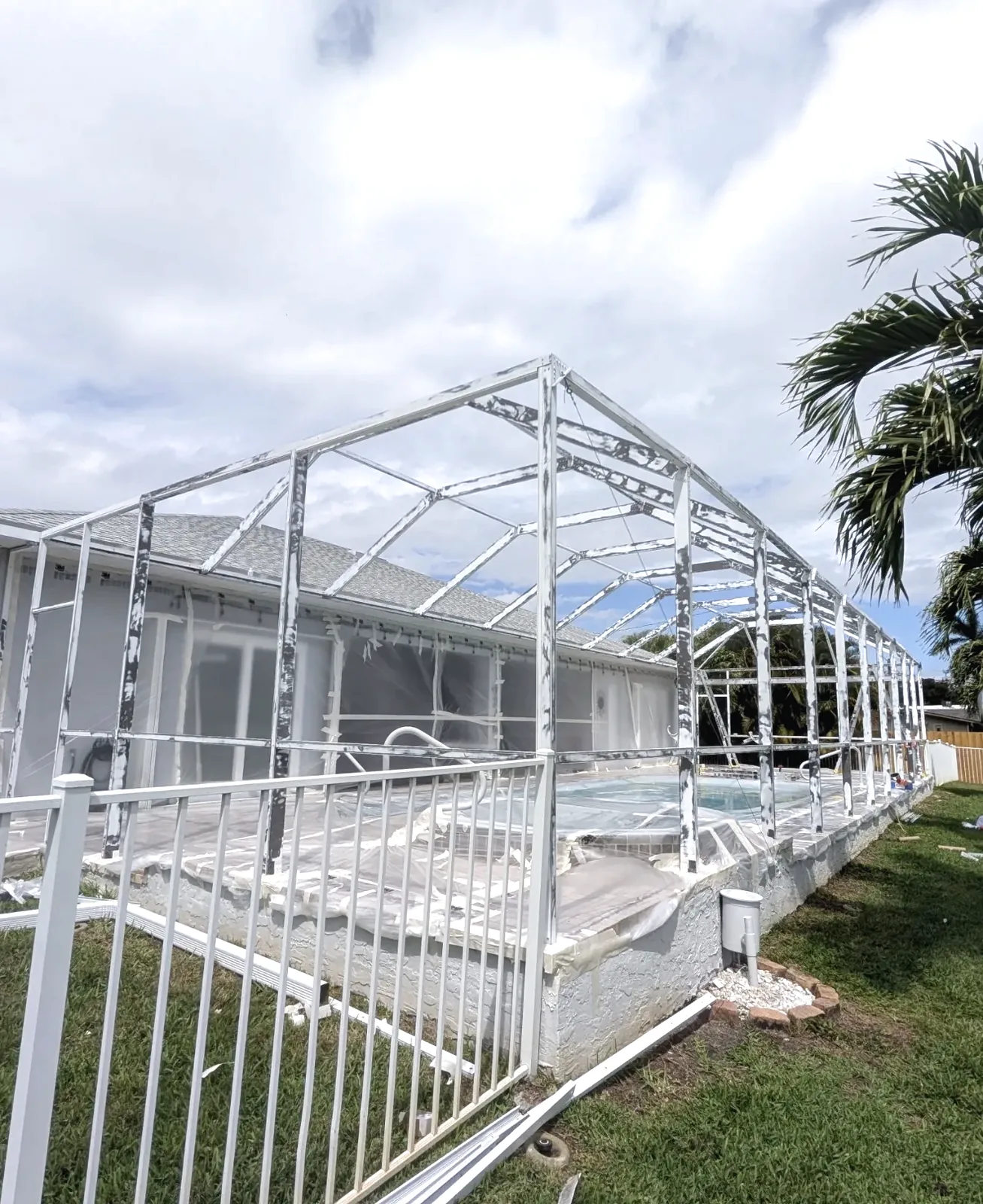 White aluminum pool cage frame stripped of screening, pool and deck covered with plastic sheeting during rescreening work.