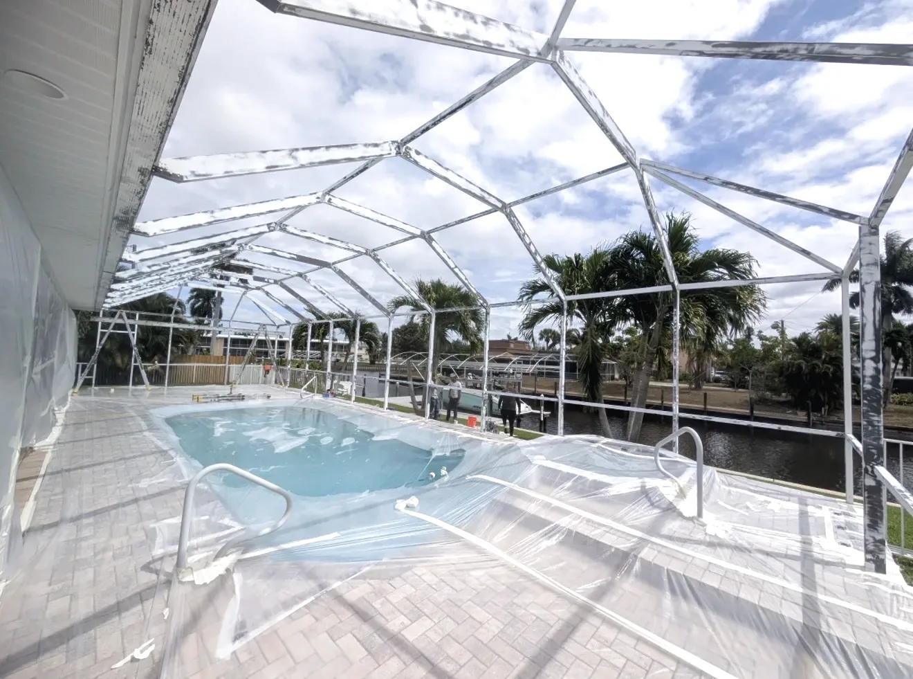 Bare white aluminum pool cage frame with no screen panels installed, plastic sheeting covering the pool and paver deck