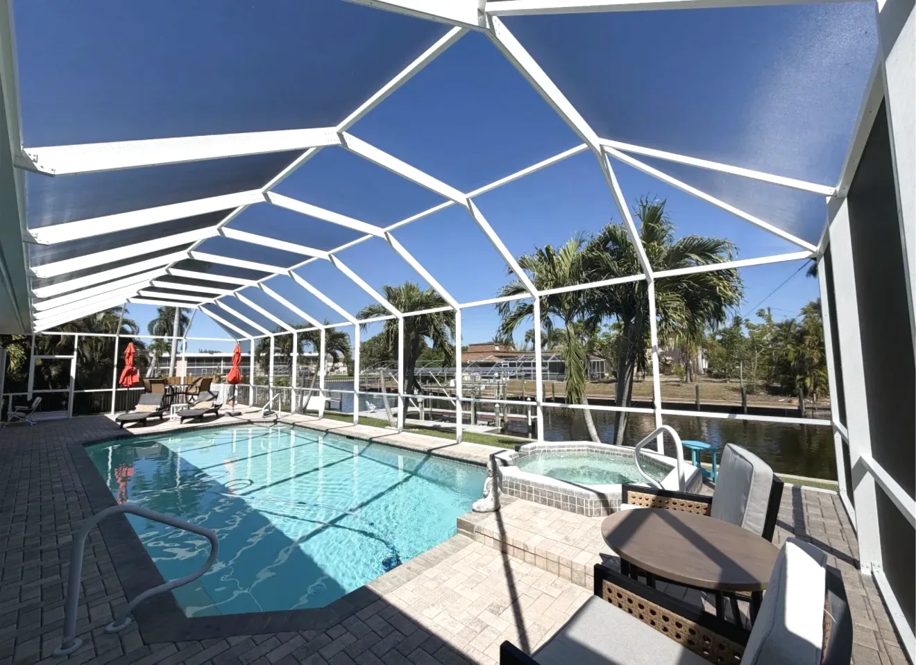 White aluminum screen enclosure over a rectangular pool and spa, paver deck with patio furniture, canal and palms behind