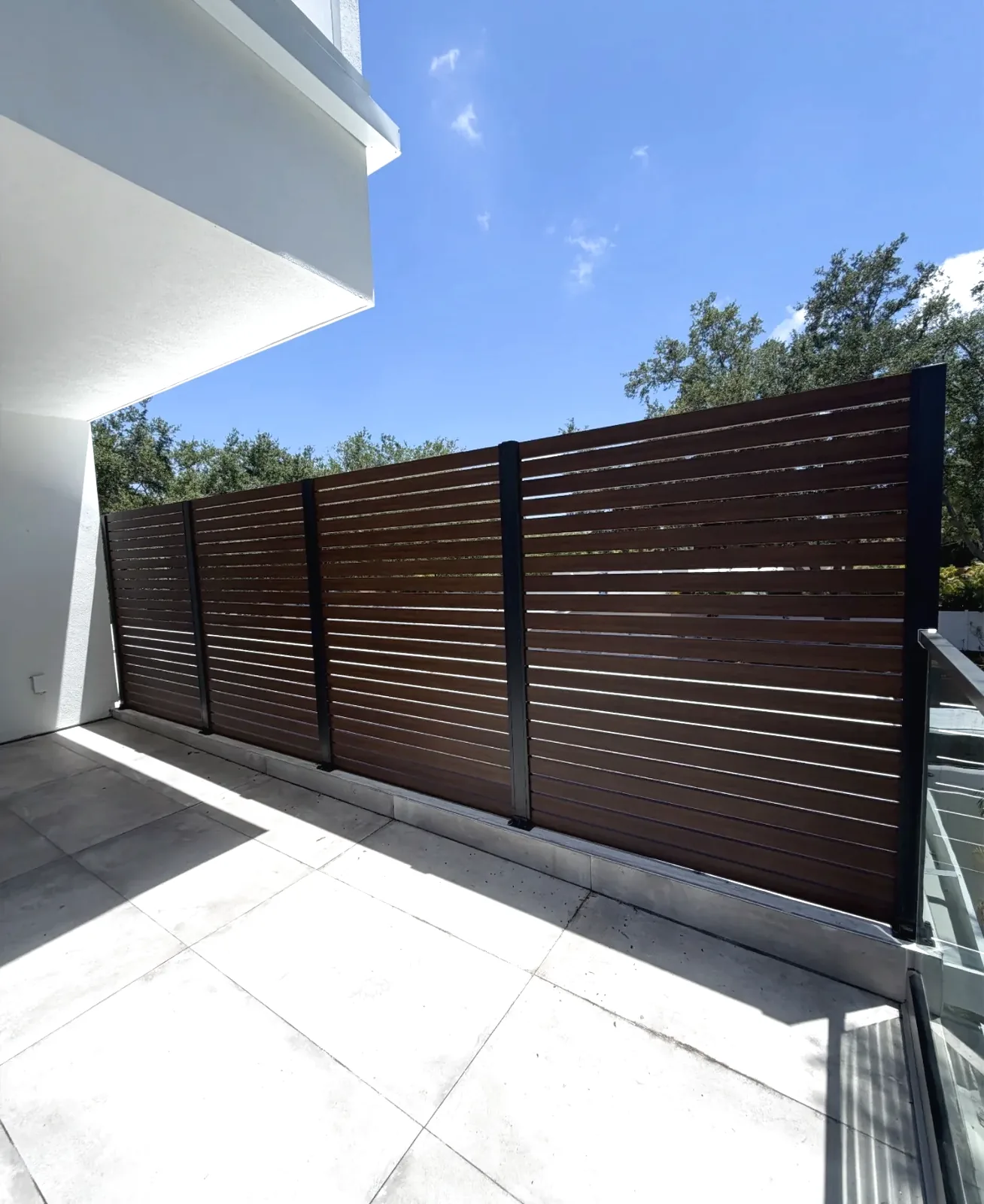 Horizontal wood-grain slat privacy screen on black posts along a tiled terrace, with a glass railing and white stucco wall