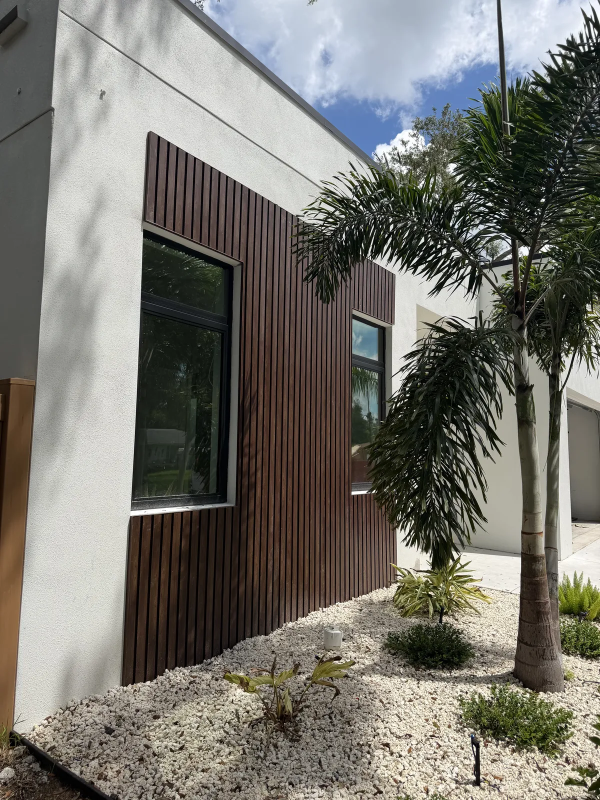 Angled view of dark woodgrain aluminum slat cladding around two black framed windows on a white stucco house, palm fronds and white rock landscaping