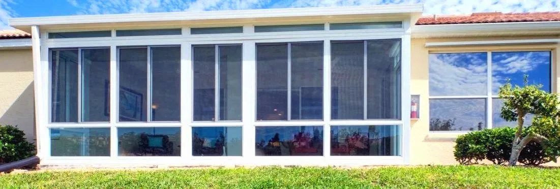 White framed room closed in with clear acrylic window panels and transom panels above, stucco house, tile roof and lawn.