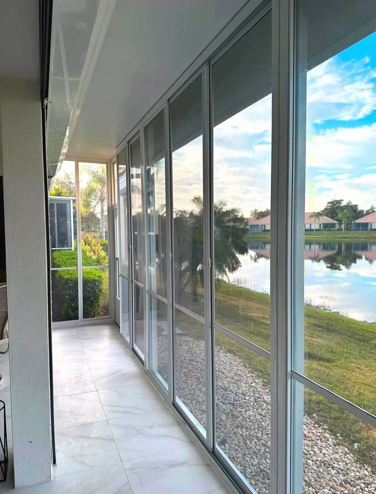 White aluminum framed acrylic sliding panels enclose a tiled lanai with river rock beds and a lake beyond