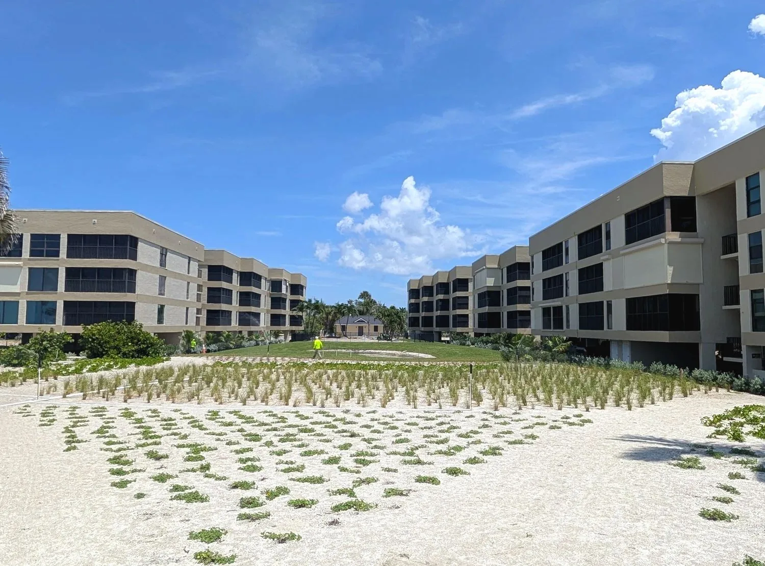 Two three-story tan condo buildings with screened lanais on every floor, dune grass and sand in the foreground, blue sky