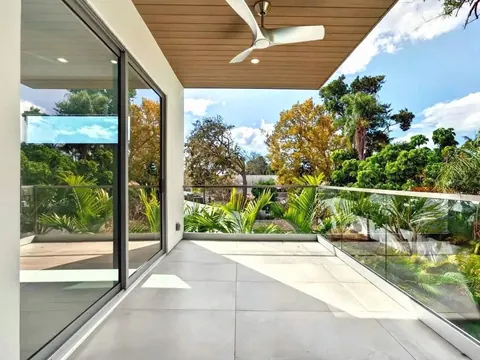 Frameless glass railing on a covered second-floor balcony with a wood ceiling and ceiling fan, trees beyond