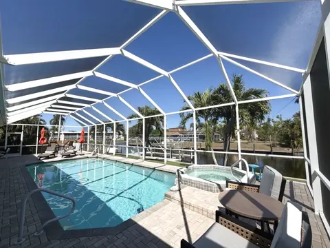 White aluminum screen enclosure over a rectangular pool and spa, paver deck with patio furniture, canal and palms behind