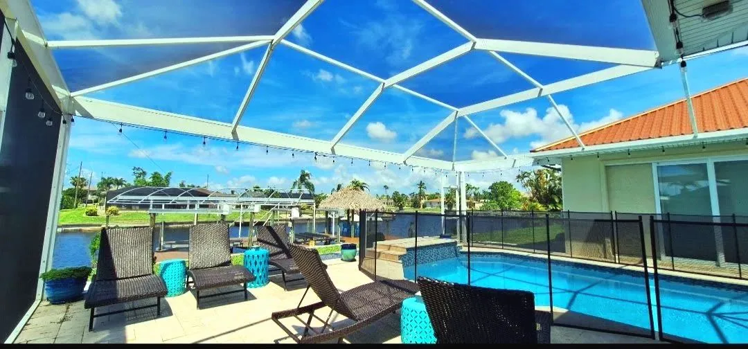 White framed pool cage over a paver deck with loungers, string lights and a canal beyond the mesh pool fence