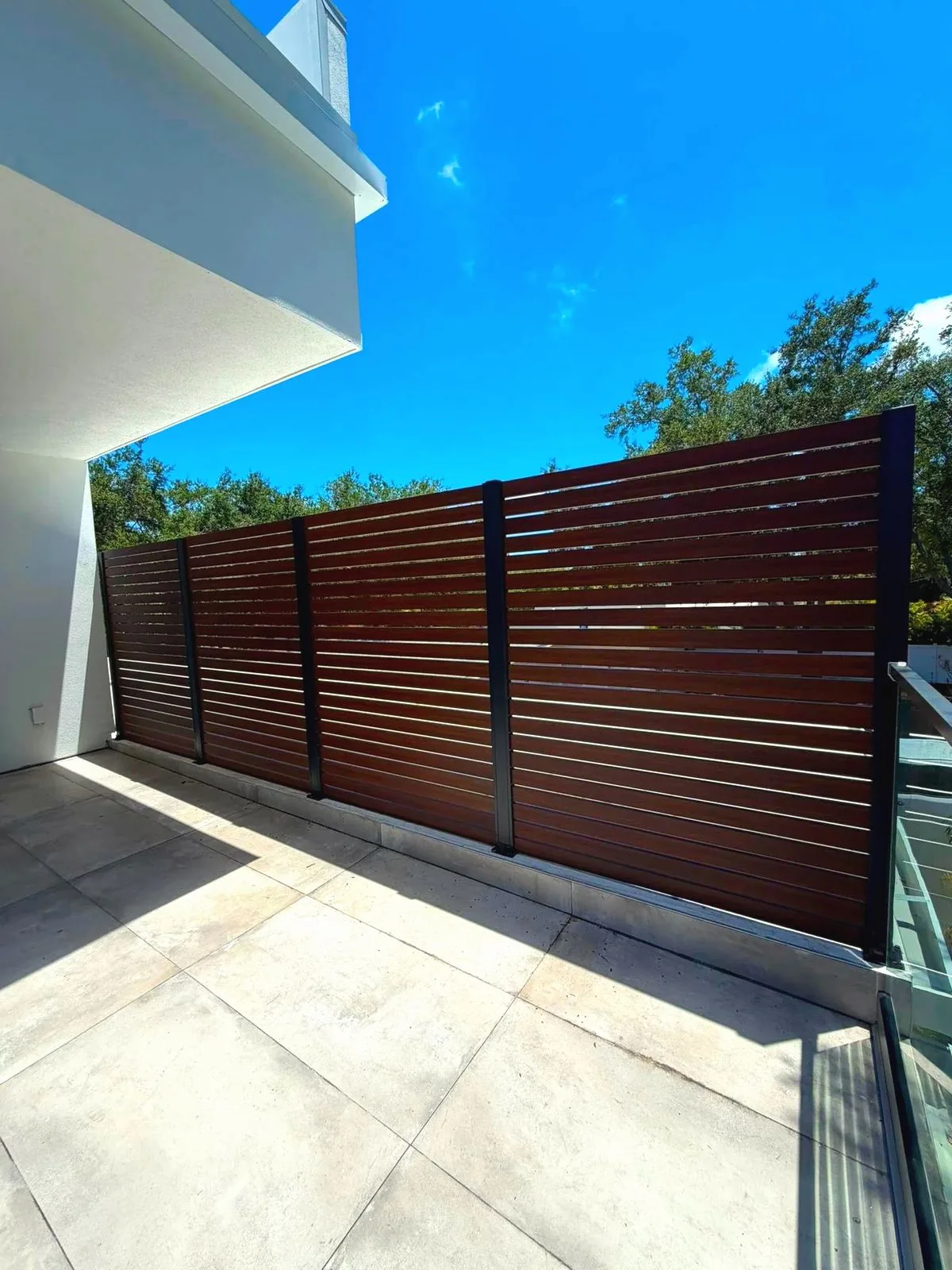 Wood-look aluminum slat privacy fence beside a white modern house, large pale pavers in the foreground, blue sky